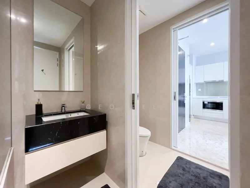 Bathroom features: single-sink vanity with black countertop, mirror above vanity, cream painted walls, tile flooring, toilet, glass shower enclosure, recessed lighting, towel rail, grey bath mat; style: modern. Room features: open doorway leading to kitchen, white cabinetry, stainless-steel appliances, light-colored flooring.