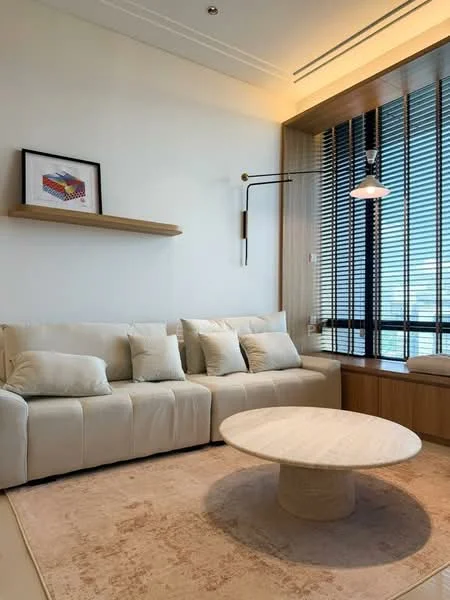Living features: 3-seater fabric sofa in cream, decorative cushions, round coffee table with marble top, area rug with neutral color, wall-mounted shelf, framed artwork on wall, floor-to-ceiling windows, horizontal wooden blinds, wooden window seat, recessed ceiling lights, wall-mounted reading lamp; style: modern; view: city.