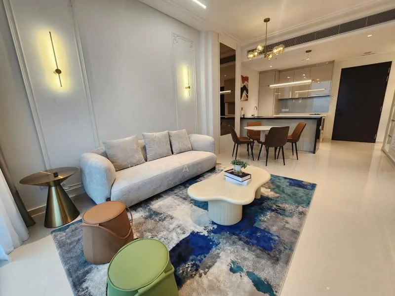 Living features: 3-seater fabric sofa in light grey, coffee table with organic shape, area rug with abstract pattern in blue and white, 2 accent stools in green and brown, wall-mounted lighting fixtures, recessed ceiling lights, open kitchen view in background, white painted walls, polished tile flooring; style: modern.