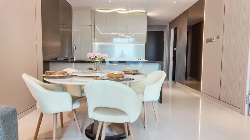 Dining features: round marble dining table, upholstered dining chairs in cream, wooden legs on chairs, decorative flower vase with pink flowers, recessed ceiling lights, white cabinetry, grey countertop, open kitchen layout, light-colored walls, polished tile flooring; style: modern. Kitchen features: white cabinetry, grey countertop, built-in appliances, recessed lighting, open layout to dining area; style: modern.
