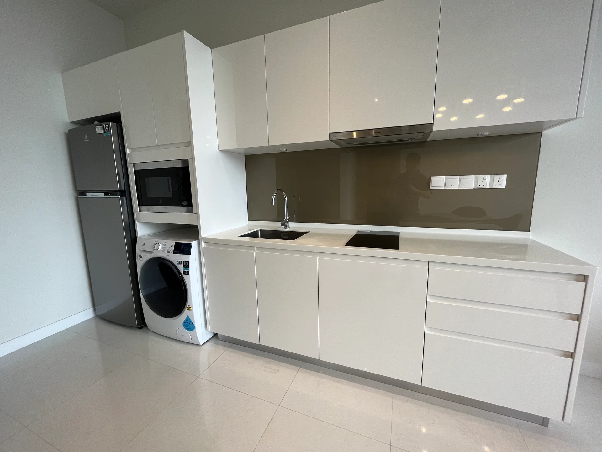 Kitchen features: white glossy cabinetry, stainless-steel refrigerator, built-in microwave, stainless-steel range hood, black glass cooktop, single-bowl stainless-steel sink, chrome pull-down faucet, white countertop, washing machine, ceramic tile flooring, recessed downlights; style: modern.