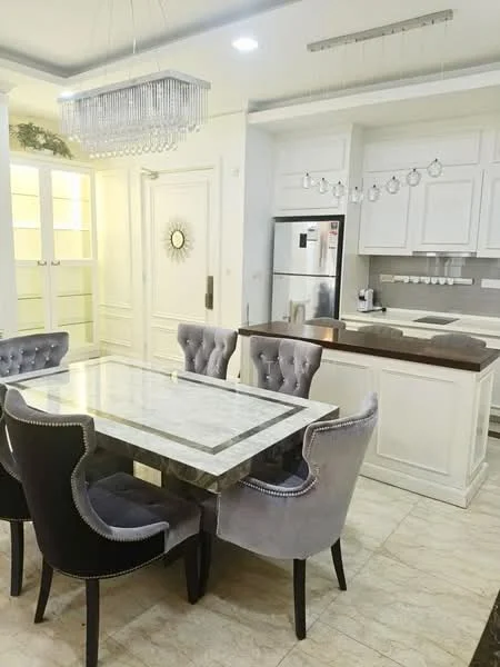 Dining features: marble dining table, 4 upholstered dining chairs in grey, chandelier with crystals, cream painted walls, tile flooring, built-in cabinetry, stainless-steel fridge, kitchen countertop with bar seating, recessed lighting, open shelving, decorative mirror, wall clock; style: modern. Kitchen features: white cabinetry, stainless-steel appliances, kitchen countertop, sink, kitchen island, built-in oven, cooktop, hanging light fixtures; style: modern.