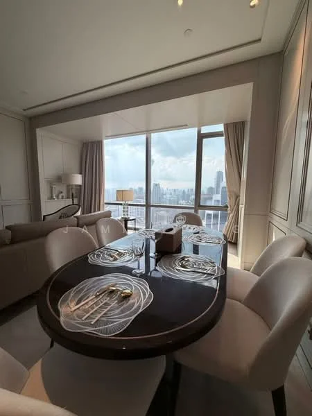 Dining features: round dining table with black surface, upholstered dining chairs in cream, set of dining plates, cutlery set, floor-to-ceiling windows, city skyline view, curtains, wall paneling, recessed lighting, decorative lamp on side table; style: modern; view: city. Living features: fabric sofa in light color, decorative cushions, coffee table, side table, floor lamp, wall paneling, recessed lighting; style: modern; view: city.