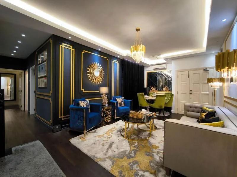Living features: dark wood flooring, blue velvet armchairs with gold accents, grey fabric sofa, gold coffee table, decorative wall mirror, gold-framed wall art, black curtains, chandelier with gold finish, recessed LED downlights, area rug with abstract pattern, gold accent decor, wall with blue paint and gold trim; style: luxury.