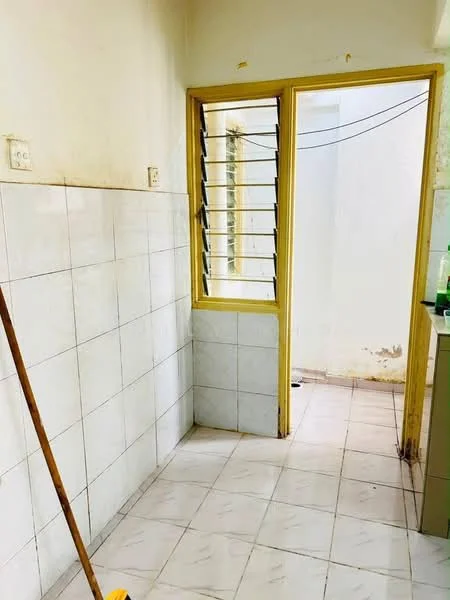 Room features: white tiled walls, cream tiled flooring, window with metal grilles, yellow painted window frame, corner sink, exposed plumbing, cleaning broom, natural light from window.