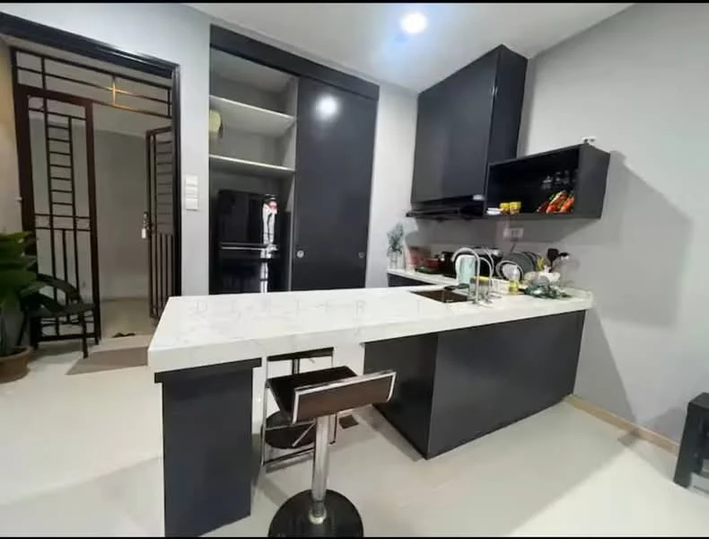 Kitchen features: black cabinetry, white quartz countertop, single-bowl stainless-steel sink, chrome pull-down faucet, built-in refrigerator, stool with chrome base, open shelving, recessed downlights, tile flooring, grey painted walls; style: modern. Room features: entrance door, decorative plant, open space leading to another room.