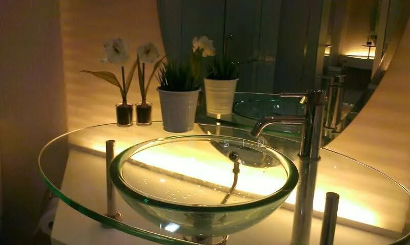 Bathroom features: glass vessel sink, chrome faucet, backlit mirror, white countertop, decorative potted plants, ambient lighting, modern design, wall-mounted storage; style: modern.