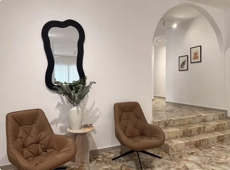 Room features: two brown upholstered armchairs, round side table, white vase with greenery, arched entryway, decorative mirror with black frame, textured flooring with marble pattern, white painted walls, recessed lighting; style: modern.
