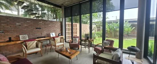 Living features: exposed brick wall, concrete ceiling, floor-to-ceiling windows, sliding glass door, wooden coffee table, 2 armchairs with wooden frames, sofa with cushions, decorative plants, area rug, wooden side table, decorative artwork, open layout, natural light, greenery view; style: modern; view: garden.
