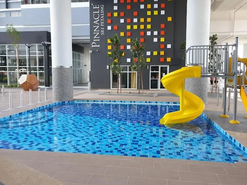 Exterior type: swimming pool deck; features: infinity pool with blue mosaic tiles, yellow water slide, playground structure, surrounding landscaped area with trees, modern building facade, grey stone cladding, colorful wall with geometric patterns, outdoor seating area; style: modern.