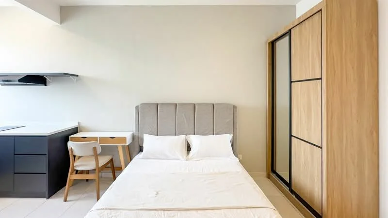 Bedroom features: queen-size bed with upholstered headboard, white bedding, built-in wardrobe with wooden sliding doors, desk with white tabletop and wooden legs, wooden dining chair, grey accent wall, light-colored tile flooring, kitchenette area with black cabinetry, white countertop; style: modern.