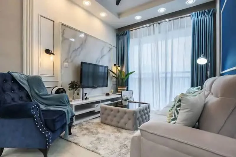 Living features: blue velvet armchair with button tufting, light grey fabric sofa, coffee table with tufted upholstery, marble accent wall, wall-mounted flat-screen TV, white media console, area rug with soft texture, potted plant, floor-to-ceiling sheer curtains, blue curtains, recessed downlights, ceiling fan; style: modern.