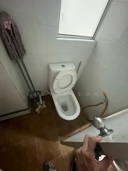 Bathroom features: white one-piece toilet, brown ceramic tile flooring, white tiled walls, window for natural light, toilet paper holder, cleaning supplies, plumbing visible, hand towel rack.