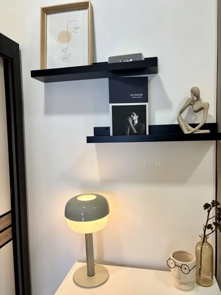 Room features: white painted walls, black floating shelves, decorative framed artwork, ceramic lamp with grey base and yellow light, decorative figurine, books on shelf, ceramic vase, dried flowers in vase; style: modern.