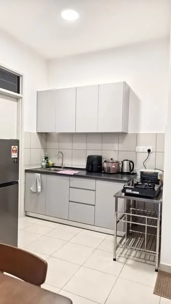 Kitchen features: grey flat-panel cabinets, black countertop, stainless-steel sink, chrome pull-down faucet, built-in oven, electric kettle, toaster, stainless-steel fridge, stainless-steel rolling cart, white tiled walls, cream tiled flooring, recessed ceiling lights; style: modern.