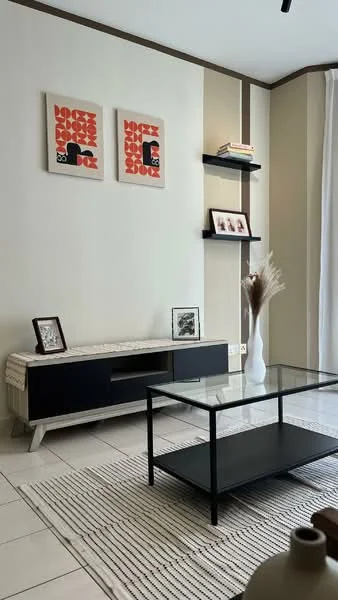 Living features: light beige painted walls, white ceiling, rectangular glass coffee table with black frame, black TV console with wooden accents, two framed artworks on the wall, open shelving unit on the wall, decorative vase on the table, area rug with striped pattern, potted decorative plant, natural light from window with sheer curtains; style: modern.