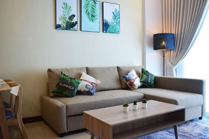 Living features: L-shape sectional sofa in beige, decorative throw pillows with tropical prints, wood coffee table, framed artwork with leaf patterns, light-colored walls, curtains with a light fabric, area rug with geometric pattern, table with four dining chairs in the background; style: modern.