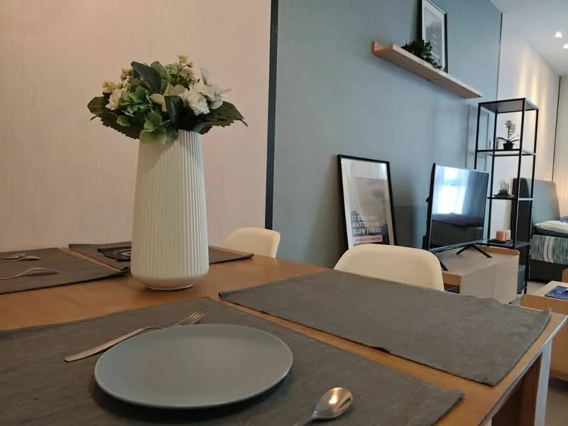 Dining features: wooden dining table, 4 dining chairs, grey placemats, ceramic plate, metal spoon, vase with flowers, light blue wall, framed artwork on wall, TV console, wall-mounted TV, shelf with decorative items; style: modern.