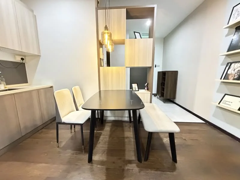 Dining features: rectangular dining table in black, 2 upholstered dining chairs in cream, bench seating in light grey fabric, wooden flooring in dark brown, pendant light fixture with warm glow, built-in cabinetry with light wood finish, open shelving with decorative items, neutral wall colour, corner of kitchen visible with light cabinetry; style: modern.