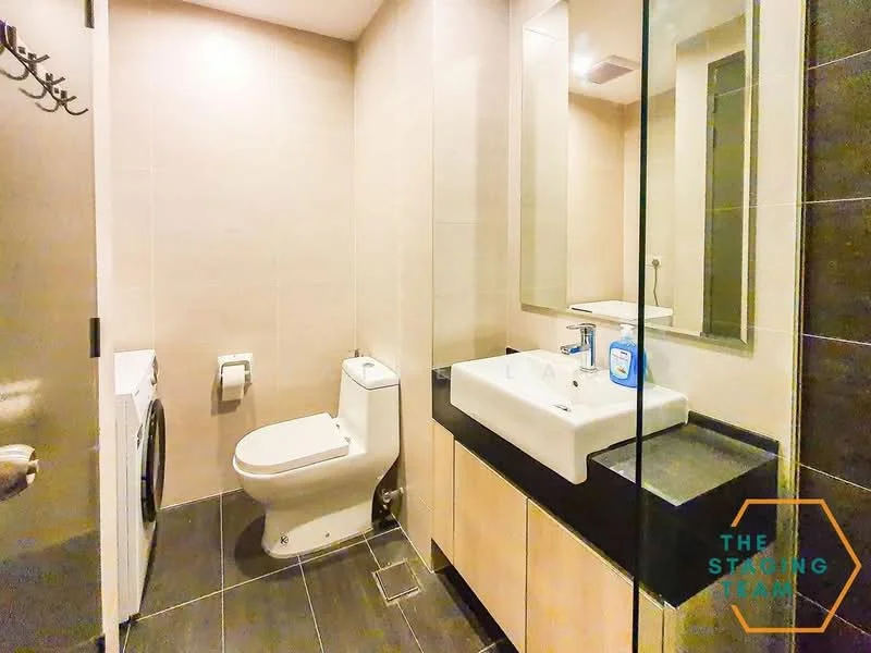 Bathroom features: wall-mounted toilet, single-sink vanity with black countertop, white ceramic sink, mirror above sink, glass shower enclosure, washing machine, grey tiled walls, black tiled flooring, recessed lighting, towel rack, chrome faucet; style: modern.