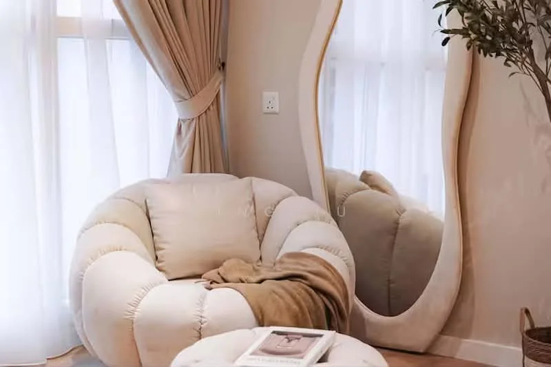 Room features: beige upholstered armchair with rounded design, tan throw blanket draped over armchair, large floor mirror with wavy frame, light-colored curtains, natural light from window, small round coffee table, decorative plant in a pot; style: modern.