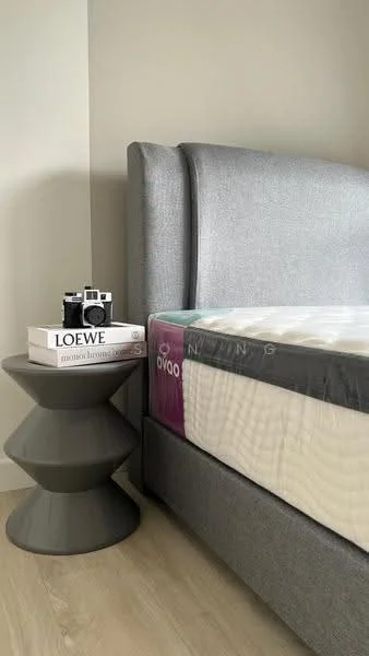 Bedroom features: king-size bed, upholstered headboard in grey, grey bedding, nightstand with unique design, stack of books on nightstand, vintage camera on nightstand, light wood flooring, neutral painted walls; style: modern.
