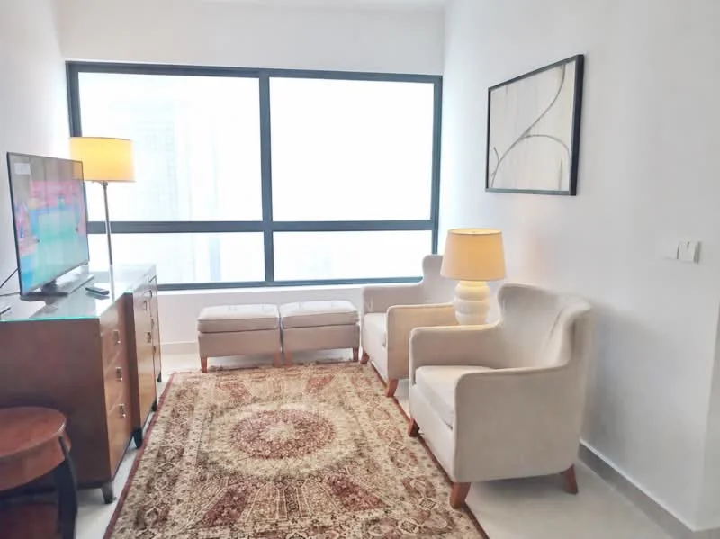 Living features: floor-to-ceiling windows, light grey walls, white ceiling, recessed downlights, 2 beige upholstered armchairs, wooden coffee table, brown wooden TV console, flat-screen TV, decorative artwork on wall, area rug with intricate pattern, table lamp with cream shade, 2 ottomans, neutral flooring; style: modern; view: city.