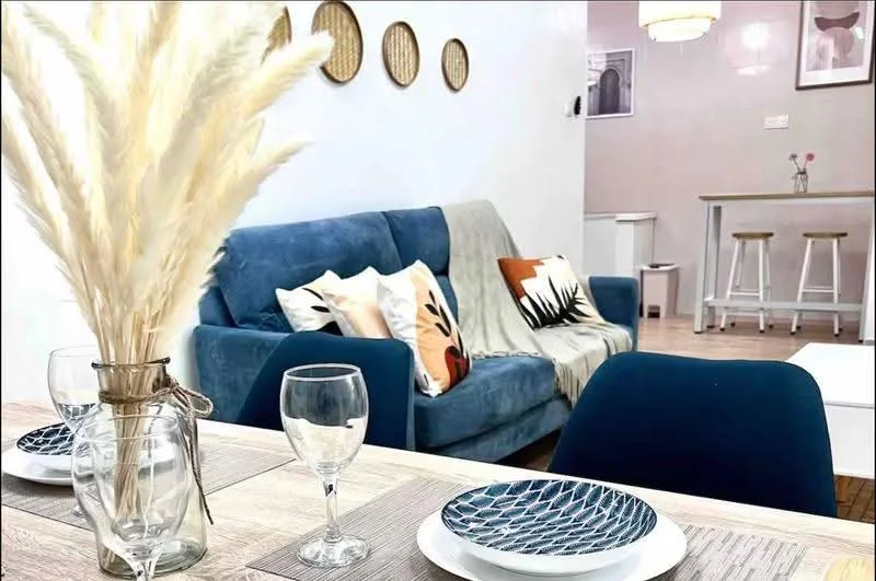 Living features: blue fabric sofa, decorative throw pillows, wooden coffee table, glassware on table, ceramic plates, wall-mounted decorative plates, light-colored walls, wooden flooring, pendant light fixture, bar counter with stools, potted plant decoration; style: modern.
