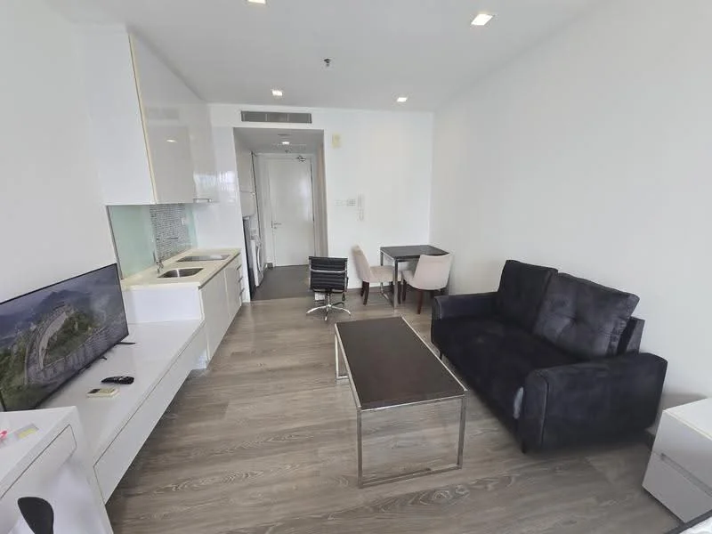 Living features: black fabric sofa, coffee table with metal legs, wall-mounted TV, TV console, dining table for four, white cabinetry, kitchenette with sink, stainless-steel appliances, wooden flooring, recessed ceiling lights, open layout, large windows, neutral wall color; style: modern.