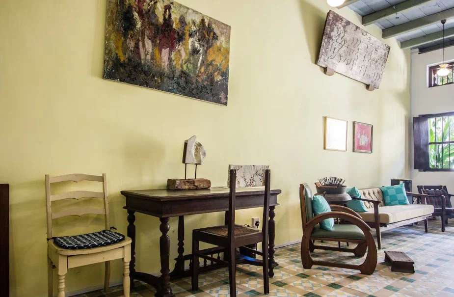 Room features: yellow painted walls, exposed ceiling beams, decorative artwork on walls, wooden table, two wooden chairs, upholstered sofa with cushions, wooden sculpture, ceramic vase, patterned tile flooring, natural light from windows; style: rustic.