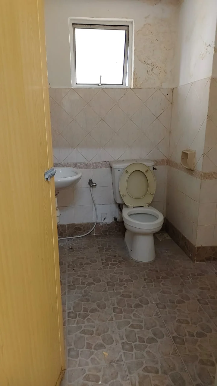 Bathroom features: white toilet, sink with chrome faucet, tile flooring with pebble pattern, beige wall tiles, white painted walls, window for natural light, toilet paper holder, shower area with no visible fixtures.
