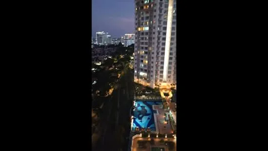 Exterior type: high-rise condominium; features: illuminated building facade, swimming pool deck, landscaped garden area, city skyline view, surrounded by urban landscape, night lighting, balconies on upper floors; style: modern.
