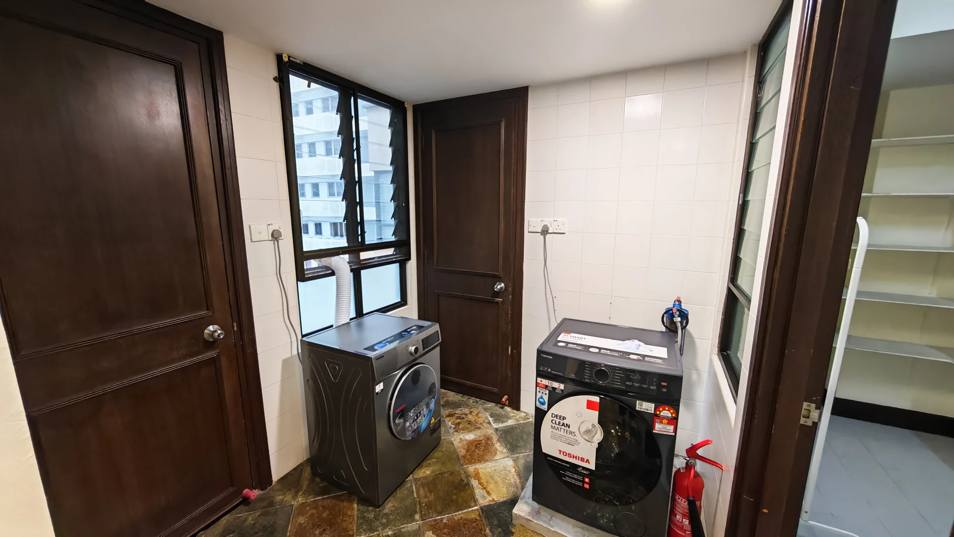 Laundry features: front-load washing machine, tumble dryer, black cabinetry, white tiled walls, slate tile flooring, two windows, wooden door, shelving unit, fire extinguisher.