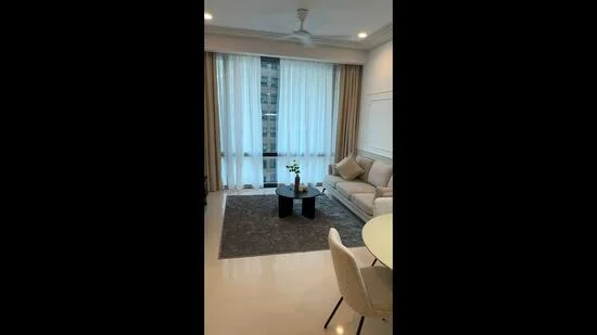 Living features: 3-seater fabric sofa in light beige, round coffee table in black, area rug with geometric pattern, floor-to-ceiling windows, sheer curtains, ceiling fan with light, decorative vase with flowers, wall-mounted artwork, recessed downlights, white painted walls, polished tile flooring; style: modern; view: city.