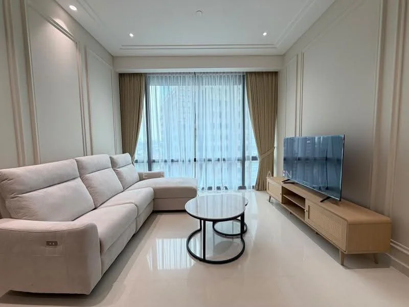 Living features: L-shape sectional sofa in light beige, round coffee table with black metal base, TV console with rattan detailing, wall-mounted flat-screen TV, floor-to-ceiling windows, sheer curtains, recessed downlights, polished marble flooring, neutral painted walls, decorative wall paneling; style: modern; view: city.