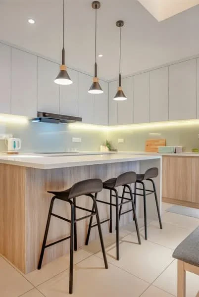 Kitchen features: white cabinetry with flat-panel design, wooden countertop, stainless-steel sink, chrome pull-down faucet, built-in cooktop, black pendant lights, backlit countertop, tile flooring, kitchen island with seating, microwave, kettle; style: modern.