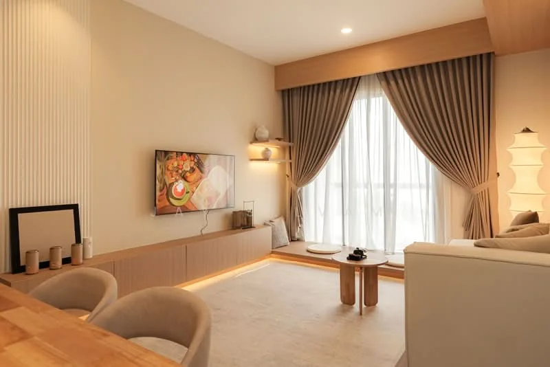 Living features: light beige painted walls, wood paneling, recessed downlights, floor-to-ceiling windows with sheer curtains, light wood cabinetry, TV mounted on wall, decorative wall shelf, neutral area rug, 2 wooden coffee tables, upholstered sofa in light color, decorative floor lamp, framed artwork, decorative objects on shelf; style: modern.