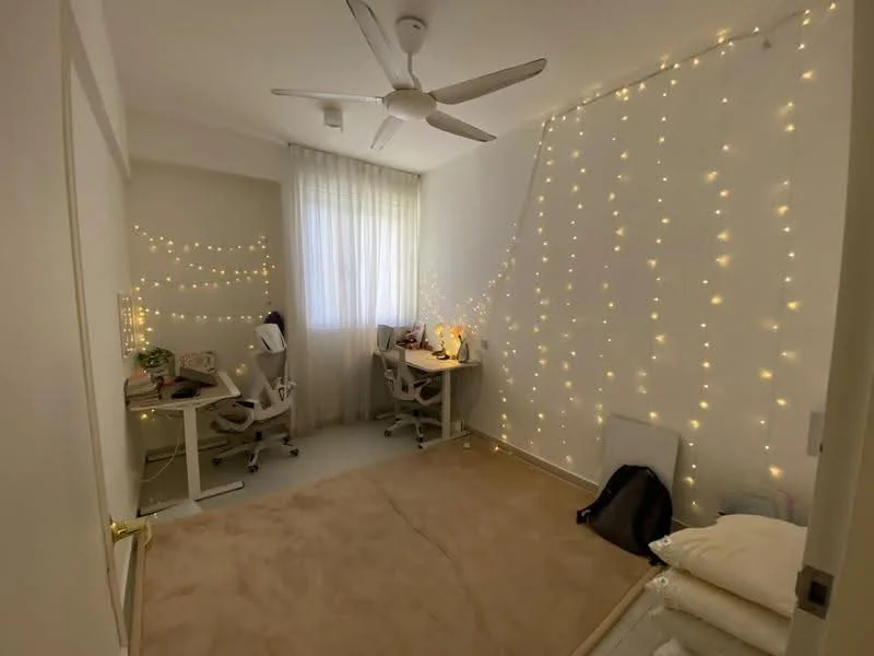 Room features: carpeted flooring, white painted walls, ceiling fan, large window with sheer curtains, decorative fairy lights on wall, two study desks with chairs, decorative items on desks, neutral color scheme; style: modern.