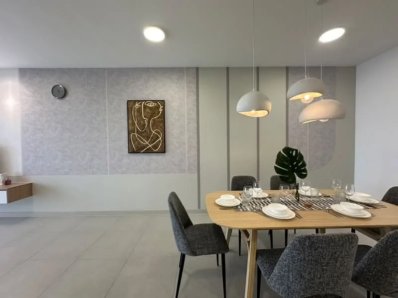 Dining features: round wooden dining table, 4 upholstered dining chairs in grey, decorative wall art, light grey accent wall, white ceiling with recessed lighting, pendant lights above the table, wall clock, neutral tile flooring; style: modern.