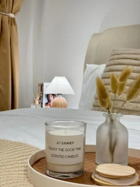 Bedroom features: queen-size bed with upholstered headboard, white bedding, decorative throw pillows, bedside lamp with pleated shade, wooden bedside table, scented candle in glass holder, decorative vase with dried flowers, woven tray, curtains in warm tone, neutral wall color, textured accent wall behind bed; style: modern.