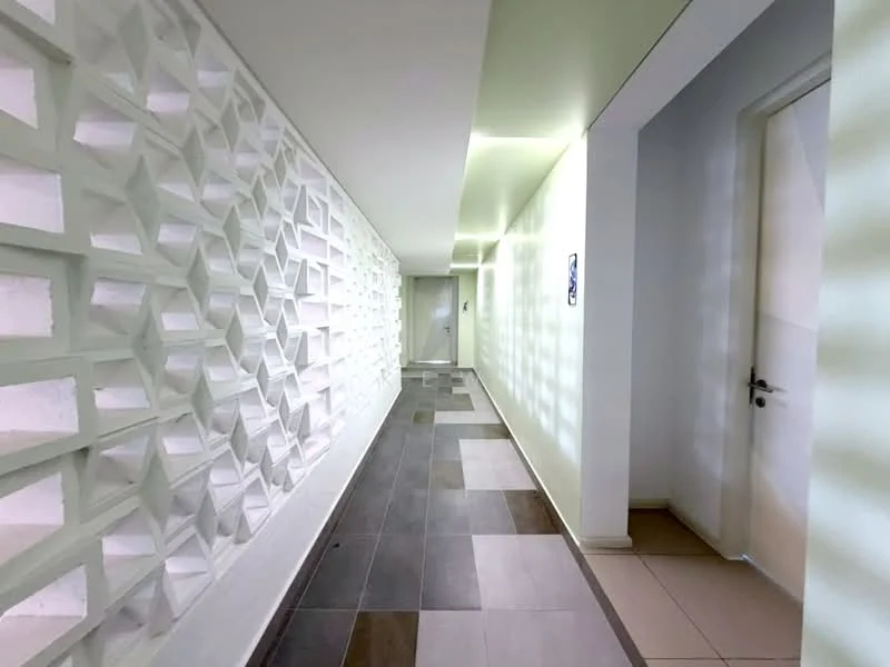 Hallway features: geometric feature wall, white painted walls, grey tiled flooring, cream tiled flooring, recessed lighting, doorway at end of hallway, smooth ceiling, decorative wall paneling; style: modern.