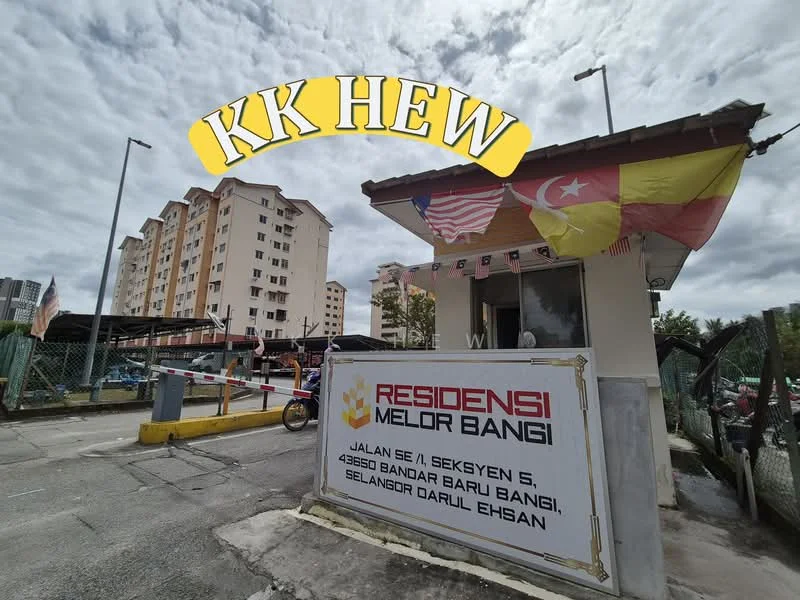 Exterior type: low-rise apartment block; features: guard house at entrance, signage with property name, Malaysian flag, state flag, surrounding fencing, parking area, multiple-storey building in background, urban context with nearby buildings; style: modern.