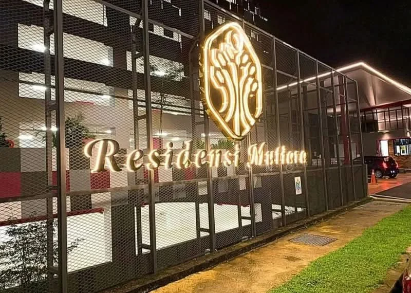 Exterior features: illuminated signage, metal mesh fencing, landscaped area with plants, paved walkway, night lighting, modern facade, parking area visible, entrance gate; style: modern.