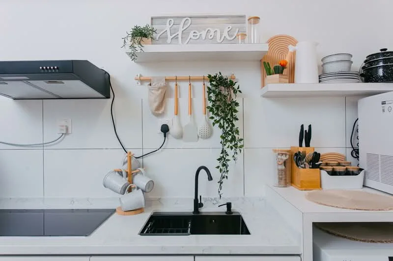 Kitchen features: black single-bowl sink, black gooseneck faucet, white marble countertop, white tiled backsplash, open shelving, wooden wall-mounted rack, hanging kitchen utensils, decorative plants, kitchen appliances, ceramic mugs, cutting board, black pots, wooden storage containers; style: modern.