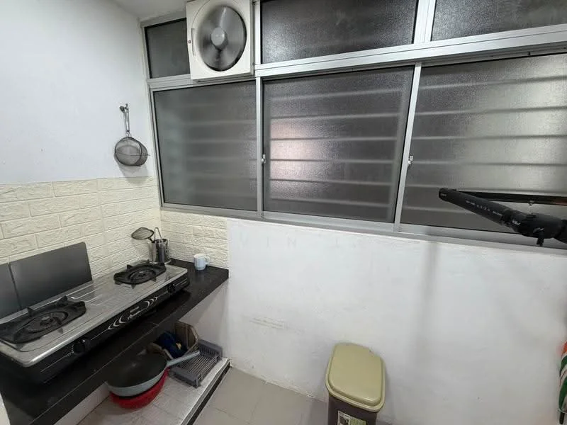 Kitchen features: black countertop, double-burner gas stove, stainless-steel kettle, white tiled walls, grey tiled flooring, window with horizontal slats, wall-mounted exhaust fan, plastic trash bin.