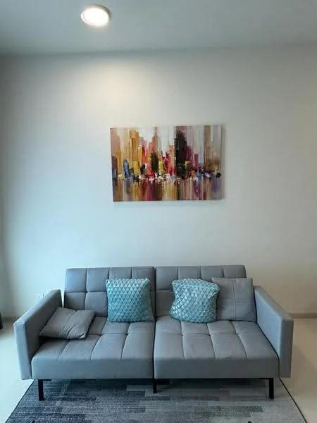 Living features: grey fabric sofa, two decorative cushions in turquoise, abstract artwork on wall, light-colored painted walls, recessed ceiling light, area rug with geometric pattern, floor-to-ceiling window, neutral tile flooring; style: modern.