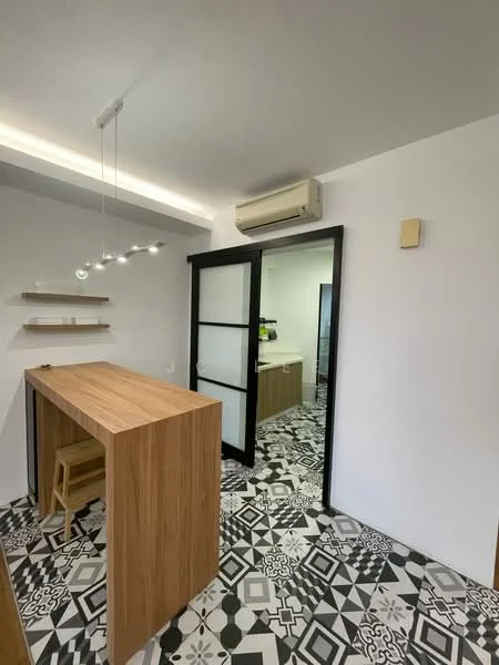 Room features: wooden bar counter, stool, black framed glass door, wall-mounted split AC, decorative pendant lighting, geometric patterned floor tiles, white painted walls, shelving unit; style: modern.