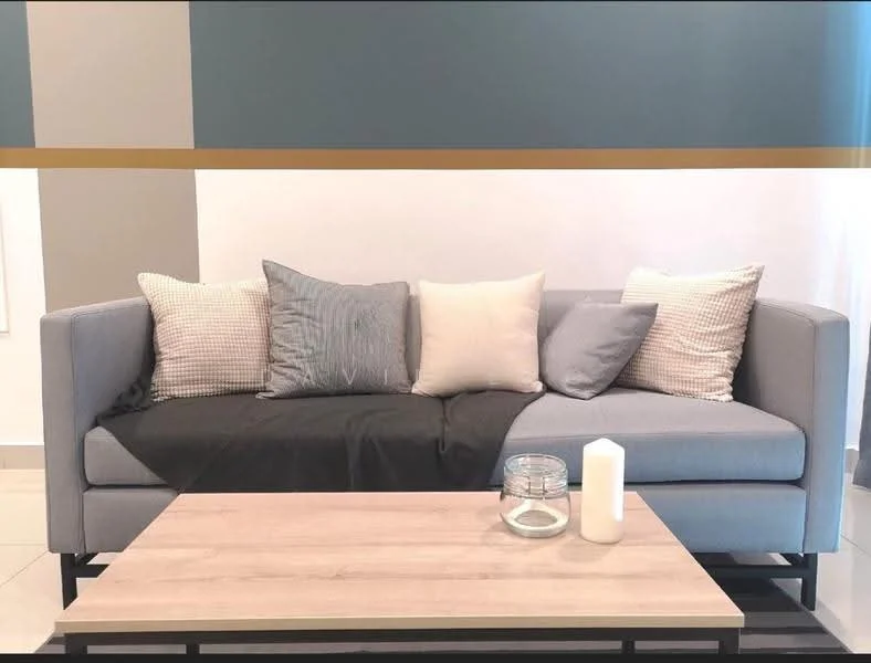 Living features: 3-seater fabric sofa in light grey, decorative throw pillows in various patterns, light wooden coffee table, glass candle holder, white pillar candle, neutral wall color, tile flooring, accent wall with gold stripe; style: modern.