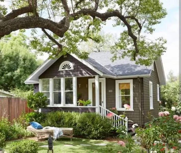 Exterior type: detached bungalow; features: wooden facade with dark brown paint, white trim around windows and doors, gabled roof with dark shingles, front porch with white railing, landscaped garden with flower beds, manicured lawn, large tree providing shade, decorative window above the front door, two outdoor seating areas, pathway leading to the entrance; style: traditional.