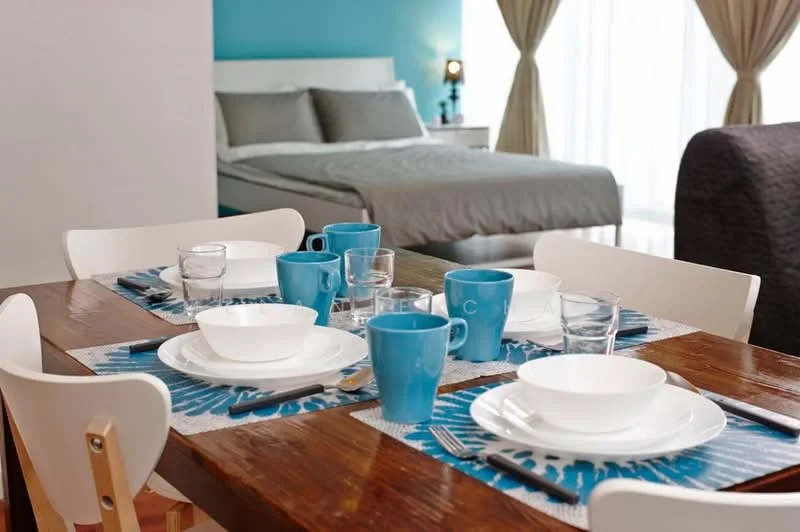 Dining features: wood dining table, white dining chairs, blue ceramic cups, clear drinking glasses, white bowls, blue table placemats, cutlery, decorative centerpiece; style: modern. Bedroom features: queen-size bed, grey bedding, white bedside table, table lamp, floor-to-ceiling windows, curtains, blue accent wall; style: modern.